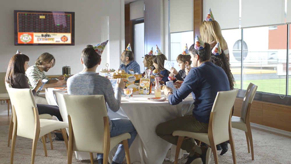 Familia y amigos celebran un cumpleaños jugando a Binvi alrededor de la mesa, con la partida en la televisión